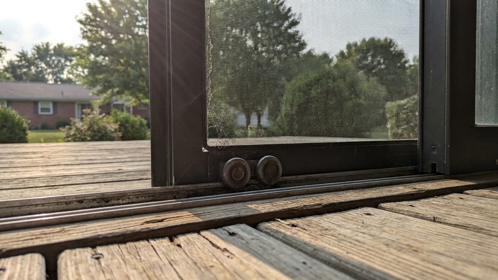 Sliding patio door screen panel partially open on a wood deck with mesh, rollers and bottom track visible in warm afternoon light